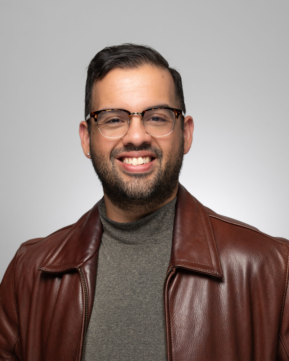 Greg Castillo smiling, wearing glasses and a brown leather jacket